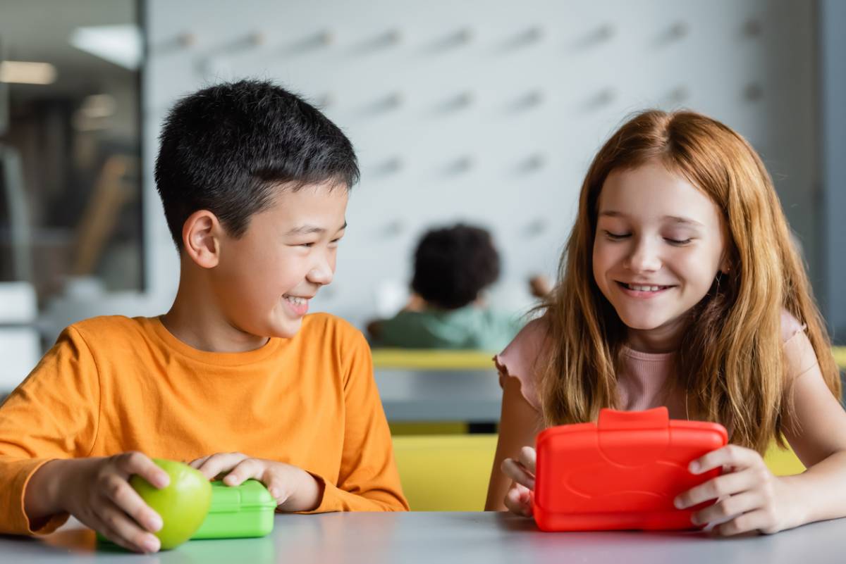 Multicultural children sit together joyfully sharing their nutritious lunches in reusable boxes.